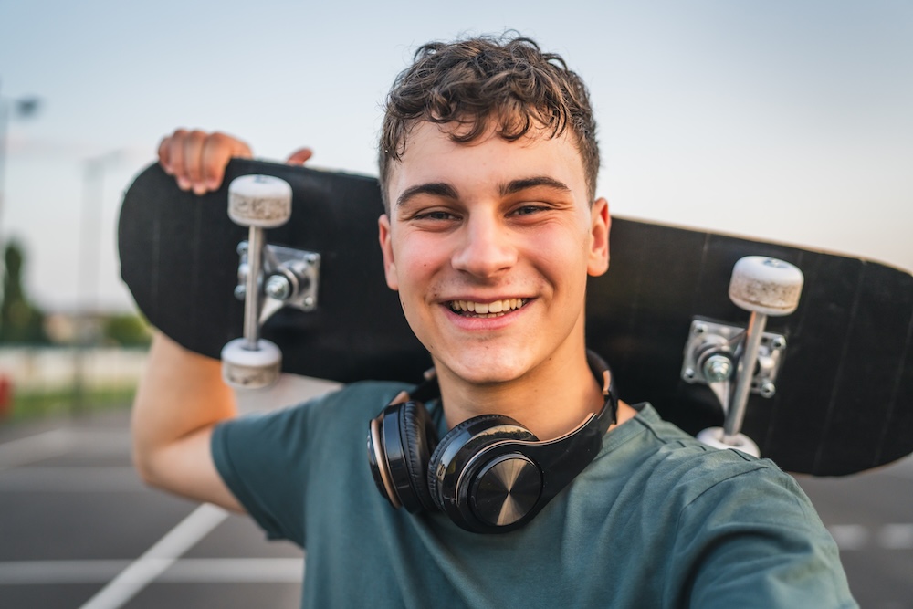 teenager boy smiling and holding a skateboard over his shoulder